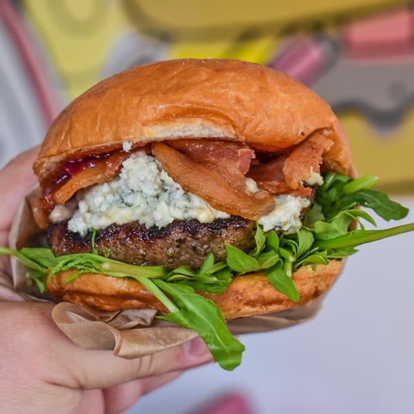 A hand holds a Sideburns burger with bacon, blue cheese, and arugula on a brioche bun.