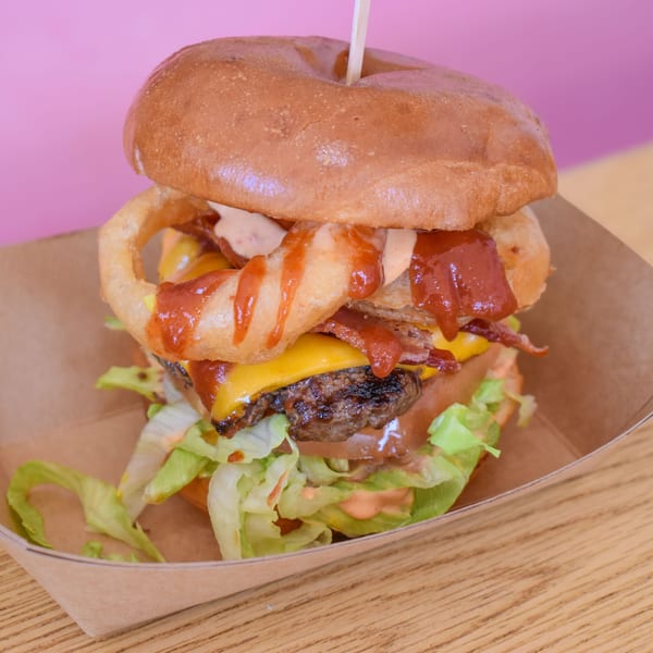 A Sideburns burger with onion rings, bacon, cheese, lettuce, and sauces in a cardboard tray.