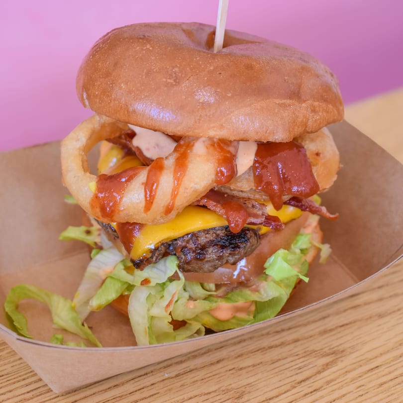 A Sideburns burger with onion rings, bacon, cheese, lettuce, and sauces in a cardboard tray.